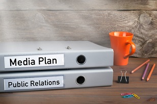 Folders on a desk labeled “media plan” and “public relations”, stacked next to pencils and a coffee mug