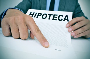 young caucasian man wearing a grey suit sitting at his office desk showing a document with the word hipoteca, mortgage loan contract in spanish, and where the signer must sign