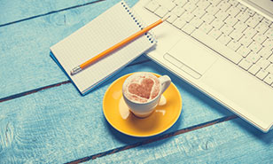 a cup of coffee with a heart drawn in the foam sitting next to a laptop and notebook